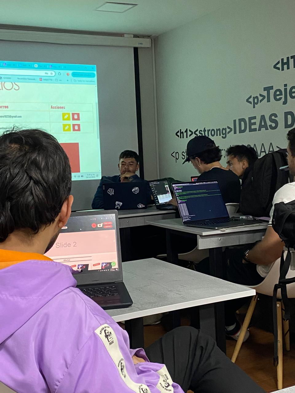 Estudiantes programando frente a sus portátiles durante una clase proyectada